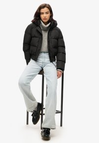 Zwarte cropped pufferjack met een capuchon, grijze gebreide trui en lichtblauwe flared jeans. Model draagt zwarte instappers en zit op een kruk.
