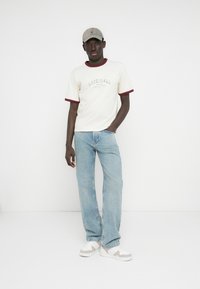 Rövid ujjú krémszínű póló bordó szegéllyel és "ORIGINALS" felirattal, világoskék egyenes szárú farmernadrággal és szürke sapkával párosítva. Fehér tornacipő teszi teljessé az öltözéket.