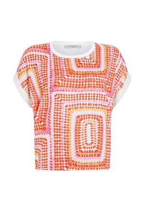 Vrouw draagt een kortemouwen top met rode, roze, gele geometrische patronen en witte manchetten, gecombineerd met zwarte jeans tegen een effen achtergrond.