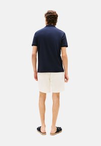 Polo shirt bleu marine, à manches courtes, de couleur unie, associé à un short clair. Sandales à enfiler teal avec un détail nœud ; design minimaliste.