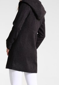 Donna che indossa un cappotto con cappuccio grigio scuro a maniche lunghe, vista da dietro, abbinato a pantaloni bianchi contro uno sfondo semplice.