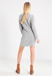 Femme aux longs cheveux blonds attachés en queue de cheval, portant une robe en maille grise texturée et des bottines marron à talons, debout devant un fond blanc.