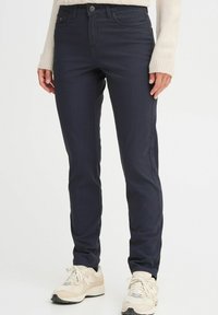Navy blue slim-fit pants with a smooth texture, featuring standard pockets and a front button closure. Paired with light sneakers.