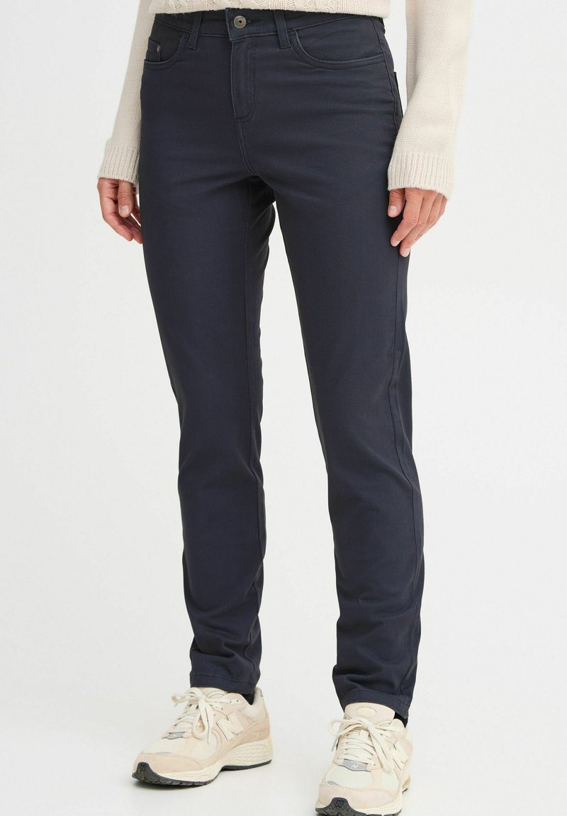 Navy blue slim-fit pants with a smooth texture, featuring standard pockets and a front button closure. Paired with light sneakers.