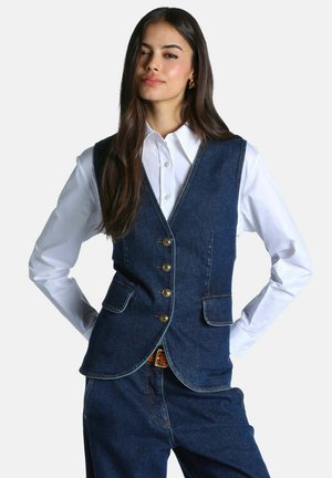 Femme aux longs cheveux foncés portant une chemise blanche boutonnée et un gilet en denim bleu foncé avec des boutons dorés, ainsi qu'un pantalon en denim assorti.