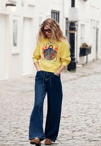 Geel sweatshirt met een "Farmer's Market Day"-beeld; gecombineerd met wijde blauwe spijkerbroek en bruine schoenen met accenten.
