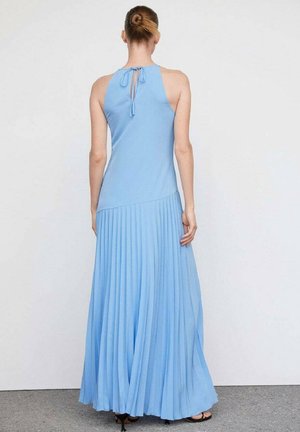 Femme avec les cheveux attachés en chignon portant une robe longue plissée sans manches bleu clair avec une fermeture par lien dans le haut du dos, debout sur un sol gris.