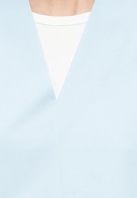 Ανοιχτό μπλε τοπ με λαιμόκοψη σε σχήμα V και λεία υφή, φορεμένο πάνω από ένα λευκό ριμπ μπλουζάκι, με καθαρό, μινιμαλιστικό σχέδιο και δομημένο σχήμα.