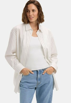 Femme aux cheveux bruns coupés au menton, portant une chemise blanche à fines rayures boutonnée par-dessus un débardeur blanc et un jean bleu clair, les mains dans les poches.