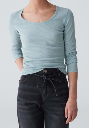 Vrouw draagt een lichtblauw langarmshirt met een ronde hals, ingestopt in een hooggetailleerde zwarte jeans met een stoffen ceintuur die is vastgeknoopt.