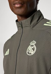 Oliivivihreä vetoketjullinen takki, jossa on korkea kaulus, vaaleanvihreät kolme raitaa ja brodannut Real Madrid -logo vasemmassa rinnassa.