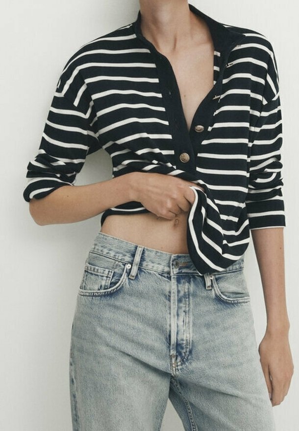 Persona che indossa una maglietta a righe bianche e nere a maniche lunghe e jeans chiari a vita alta, sollevando la maglietta per rivelare la parte centrale del corpo.