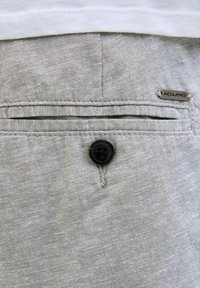 Light gray fabric, featuring a single black button and a small metal label with brand name. Simple pocket design, textured surface.