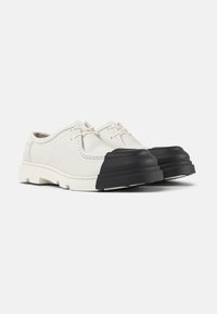 Chaussures blanches en cuir avec des bouts en caoutchouc noir, des lacets et une semelle texturée. Le design présente des lignes épurées et des matériaux contrastants.
