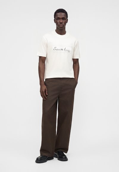 Άνδρας που φοράει λευκό t-shirt Emporio Armani, καφέ παντελόνι με φαρδιά πόδια και μαύρα παπούτσια στέκεται μπροστά σε απλό ανοιχτό φόντο.