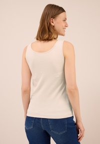 Donna con un top senza maniche beige chiaro e jeans blu, in piedi con le spalle rivolte verso l'osservatore, sorridendo, su uno sfondo beige uniforme.