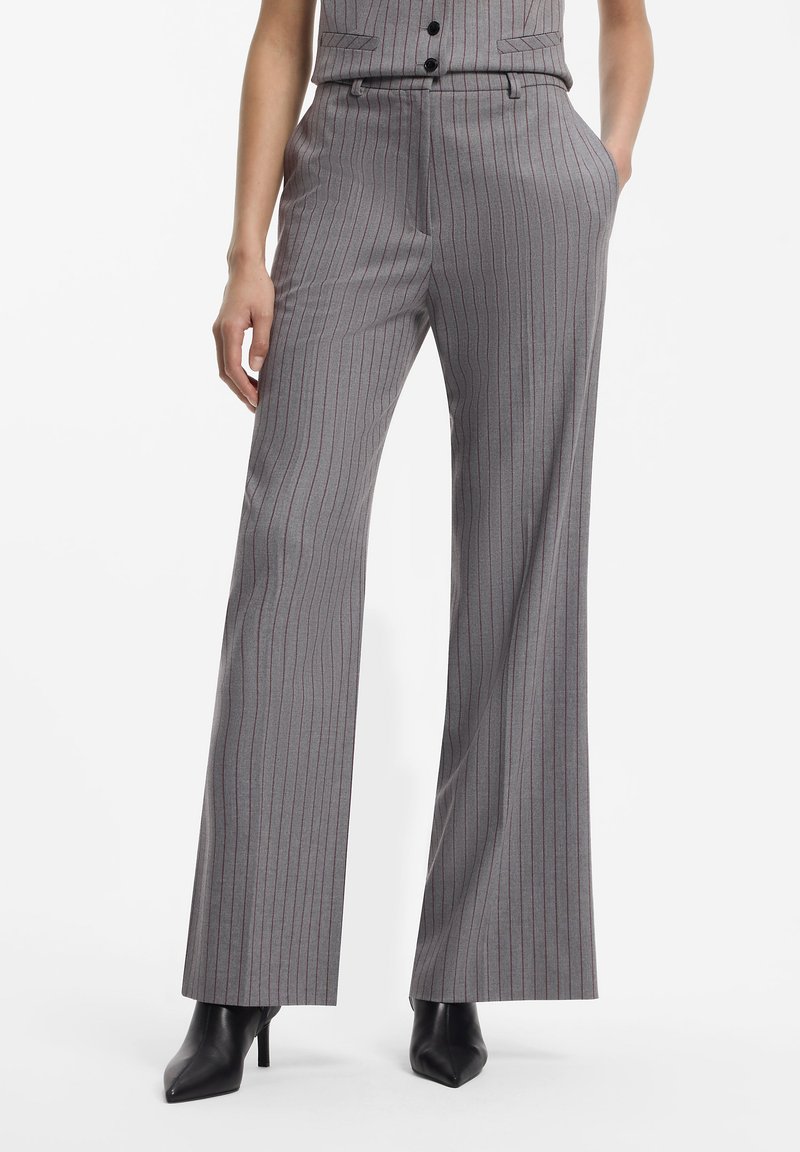 Femme portant un pantalon large gris à rayures fines avec des bottes noires à talons aiguilles pointues, une main dans la poche, sur fond blanc.