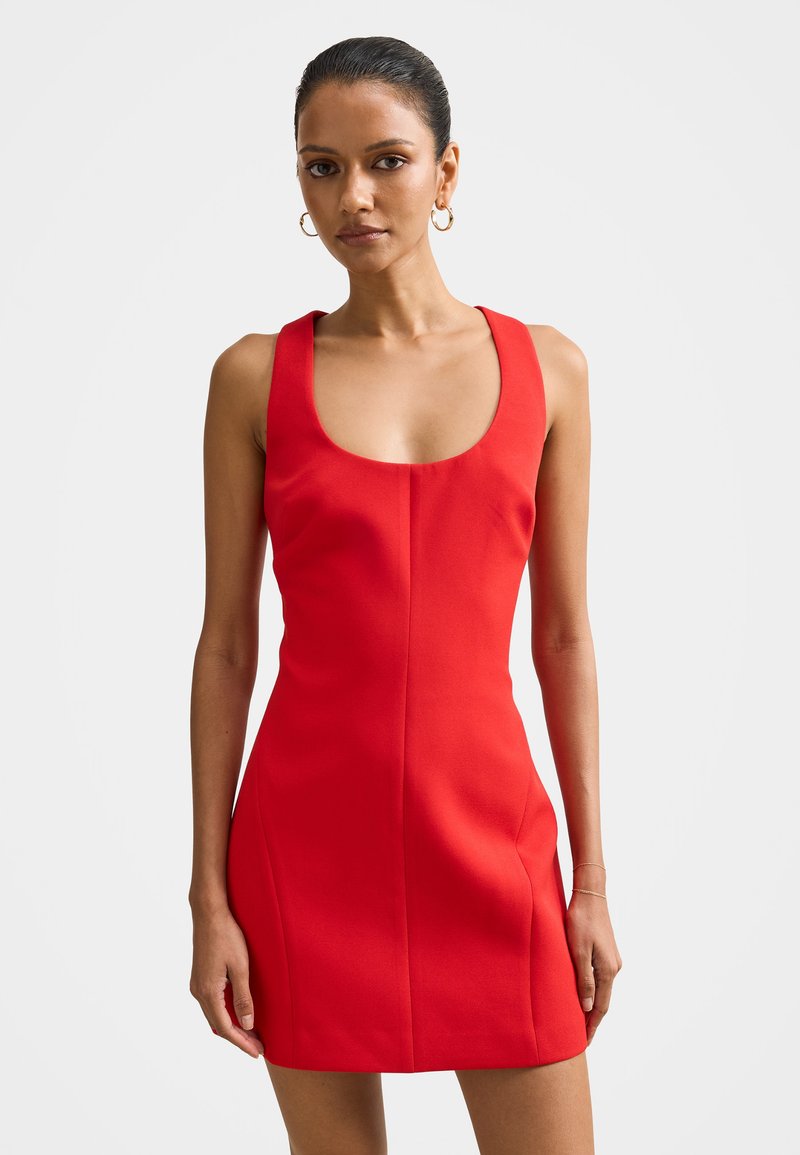 Tumejuustega naine punases varrukatuta kehailevas kleidis, millel on ümardatud kaelus, seisab lihtsa heleda tausta ees.