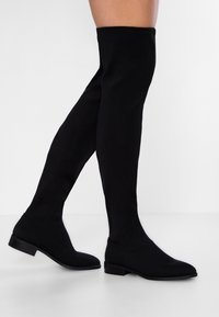 Botas negras hasta arriba de la rodilla, hechas de tela suave, con punta afilada, tacón plano y diseño sin costuras que se extiende hasta la mitad del muslo.