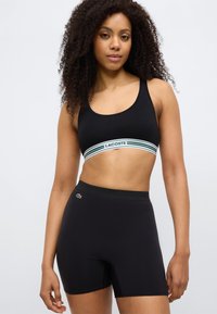 Brassière de sport noire avec bande rayée verte et blanche arborant le logo "LACOSTE" ; associée à un short taille haute noir avec un petit logo.