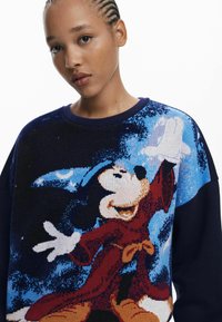 Strickpullover mit einem blauen und marineblauen Design, das eine Grafik eines Charakters in einem roten Gewand zeigt. Strukturierter Stoff und gerippter Halsausschnitt.