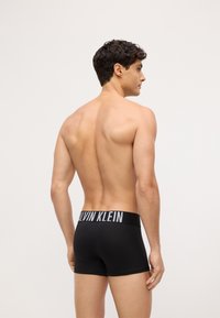 Crne bokserice s širokim elastičnim pojasom na kojem piše "CALVIN KLEIN". Glatka tkanina s uskim dizajnom, prikazuje stražnji pogled.