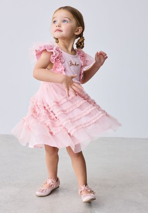 Mladé dievča s vrkočmi sa točí v vrstvených ružových šatách a zodpovedajúcich kvetinových topánkach na jednoduchom svetlom pozadí.