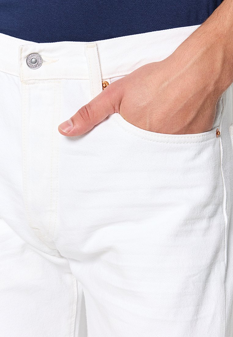 Weiße Denim-Jeans mit einem Knopfverschluss und einer Seitentasche, die eine glatte Textur und eine maßgeschneiderte Passform zeigen. Dezente Steppnähte sind sichtbar.