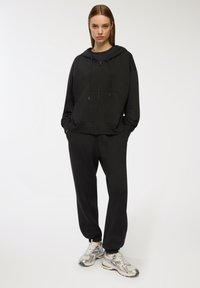 Czarna bluza z kapturem na zamek błyskawiczny z kangurzą kieszenią, w połączeniu z dopasowanymi dresami i metalicznymi butami sportowymi. Gładki materiał i luźny krój.