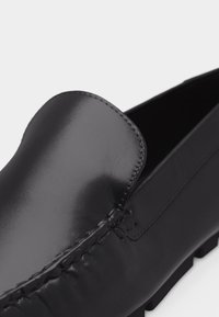 Mocassin en cuir noir avec une finition polie lisse et des coutures détaillées le long de l'avant et des côtés, présenté en gros plan.