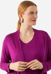 Fuchsia-langärmelige Strickjacke über einem passenden kurzärmeligen Oberteil mit einer glatten Textur und offenem Design. Es werden schwarze rechteckige Ohrringe getragen.