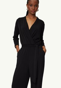 Czarny kombinezon z długim rękawem, dekoltem w serek i paskiem w talii. Posiada boczne kieszenie oraz gładki, teksturowany materiał.