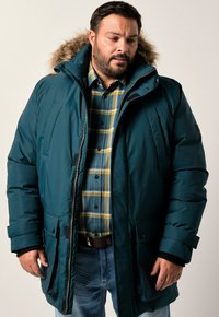 Giacca puffa turchese con cappuccio foderato in pelliccia, cerniera frontale e tasche con bottoni. Indossata sopra una camicia a quadri blu e gialla. Tessuto morbido.