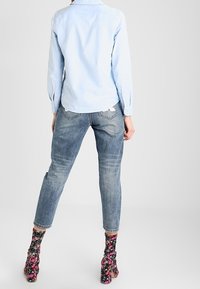 Person som bär en ljusblå skjorta, avkortade blå jeans och svarta blommiga högklackade stövlar, står med ryggen vänd framåt.