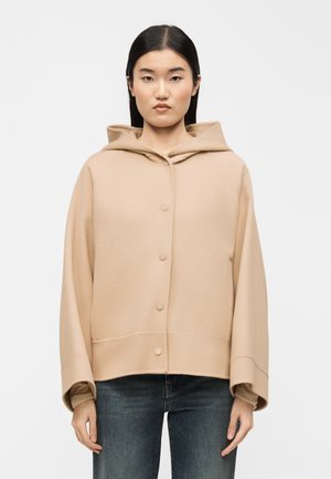 Kobieta o prostych czarnych włosach nosi beżowy, zapinany na guziki płaszcz z kapturem oraz niebieskie dżinsy, stoi na tle jasnego, jednolitego tła.