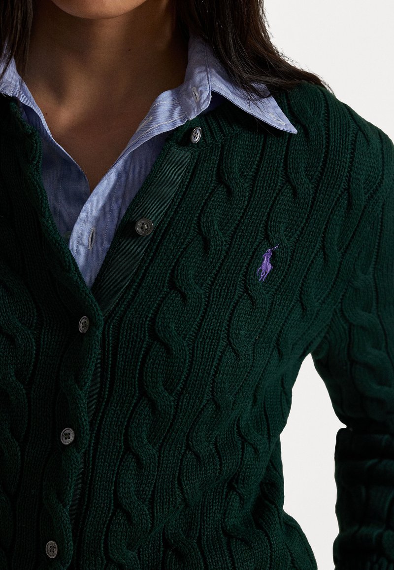 Mørkegrøn kabelstrikket cardigan med V-udskæring, der har sølvknapper og et broderet logo i lilla på venstre side. Båret over en lyseblå skjorte.