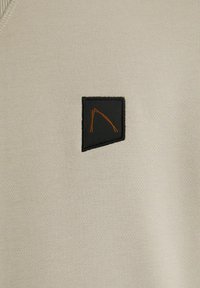 Maglione beige chiaro realizzato in morbido tessuto, con una toppa nera che presenta un motivo geometrico arancione. Texture liscia e design semplice e moderno.