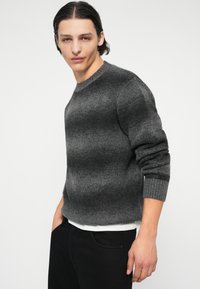 Ένα γκρι πλεκτό πουλόβερ με ριγέ σχέδιο σε διάφορες αποχρώσεις. Έχει στρογγυλή λαιμόκοψη και ριμπ μανσέτες, συνδυασμένο με μαύρο παντελόνι.