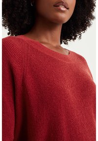 Punainen neulepaita, jossa on ribbitekstuuri, leveä kaula-aukko ja laskettu olkasauma. Kangas on pehmeä ja näyttää lämpimältä.