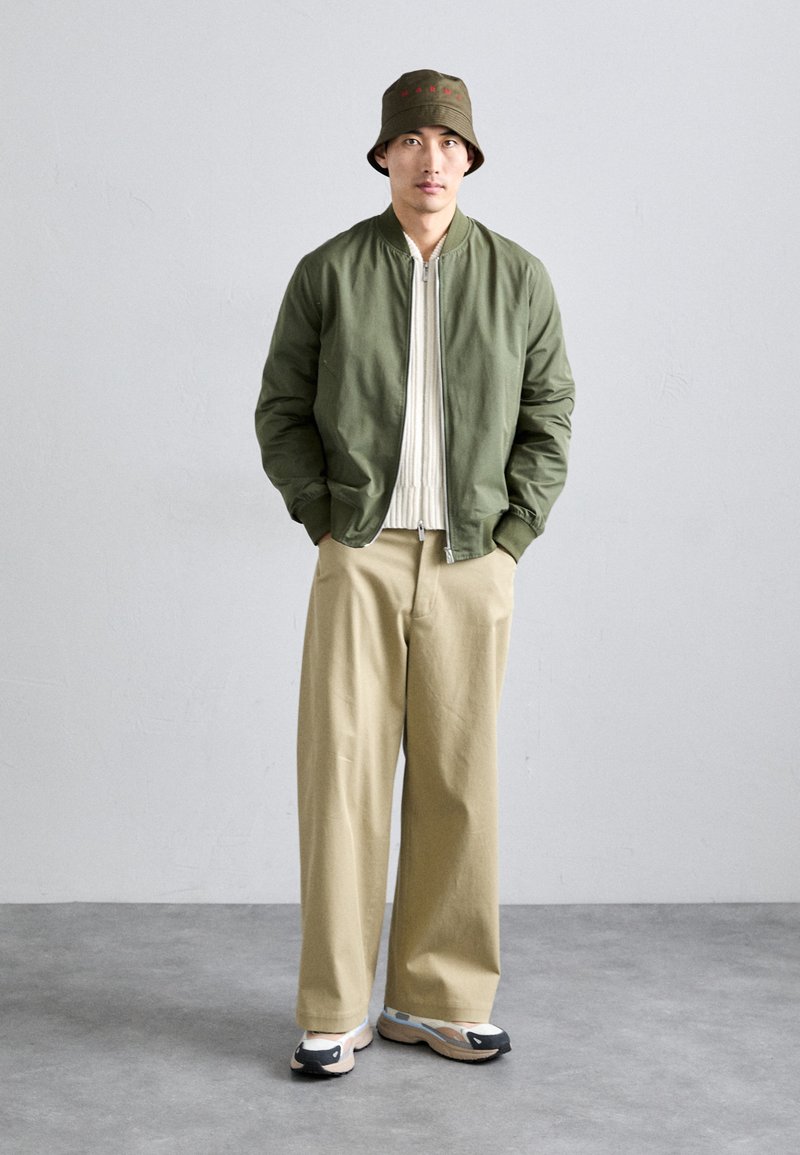 Grüne Bomberjacke über einem weißen Rippenhemd, beigere weit geschnittene Hose und schwarze/graue Sneaker. Ein grüner Bucket Hat mit rotem Text.