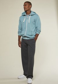 Helle blaue Zip-Hoodie mit weißen Kordeln, kombiniert mit dunkelgrauen Jogginghosen und weißen Sneakern, präsentiert an einem Model.