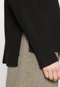 Svart ribbad tröja med avslappnad passform, med en sidofick. B ärs över ett strukturerat, ljusgrått tyg. Slät textur och enkel design.