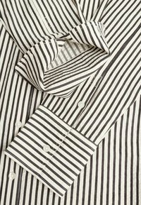 Manche de chemise blanche à rayures verticales noires, montrant un poignet plié et trois boutons blancs sur la patte de boutonnage et le poignet.