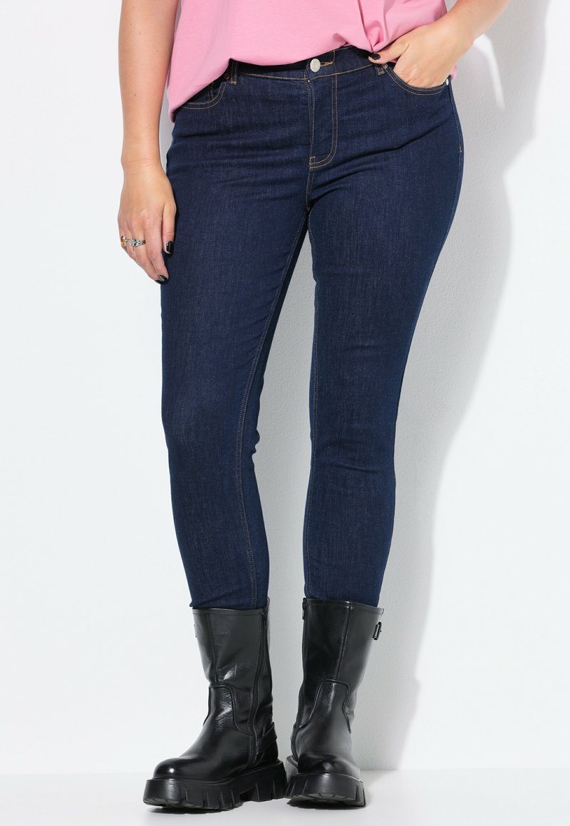 Jeans ajustados de mezclilla azul oscuro, con cintura de altura media, bolsillos laterales y costuras contrastantes. Combinados con botas negras de tobillo.