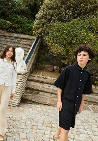 Twee kinderen lopen op stenen trappen. De ene draagt een witte denim jas en beige broek. De ander heeft een zwart overhemd met knopen en shorts aan. Groene bladeren omringen hen.