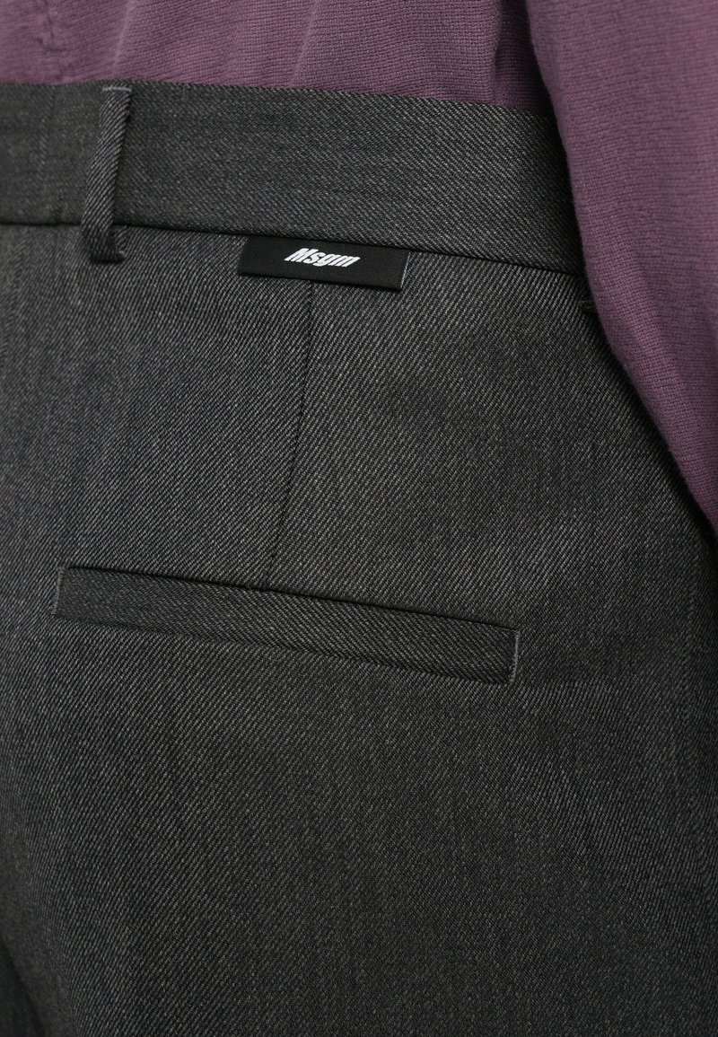 Gros plan sur un pantalon tailleur gris foncé avec une poche passepoilée arrière et une étiquette de marque noire, associé à un haut texturé violet.