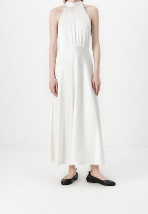 Biała maxi sukienka z halterowym dekoltem, marszczonym pasem i zwiewną spódnicą, wykonana z lekkiego materiału, w połączeniu z czarnymi balerinami.