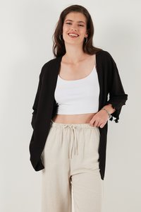 Cárdigan negro con acentos de flecos, usado sobre una camiseta sin mangas blanca y pantalones sueltos beige claros. Textura suave y diseño de apertura en el frente.