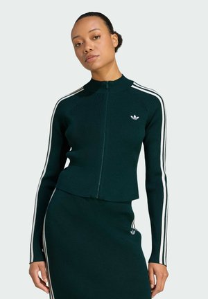 Green ribbed zip-up jacket with white side stripes, featuring a high collar and an embroidered logo on the chest, paired with a matching skirt.