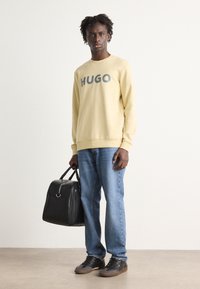 Gul genser med "HUGO" trykt i grått, blå jeans, svart veske og mørke joggesko. Glatt stoff med avslappet passform og diskré design.